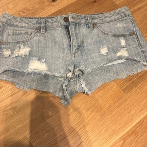 Ljusblåa lågmidjade  jeansshorts med slitningar - Säljer ett par ljusblå jeansshorts med flera slitningar för en cool, avslappnad vibe. Klassisk femficksmodell med knapp och dragkedja framtill. Perfekta för varma dagar och festivalhäng.
