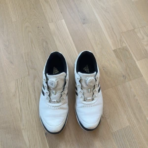 Vita golfskor från Adidas med BOA-snörning - Säljer ett par vita golfskor från Adidas med svarta ränder på sidorna och BOA-snörningssystem för enkel justering. Skorna har perforerad ovandel för ventilation och en robust sula med grepp för golfbanan. Tillverkade i skinn och syntetmaterial, riktigt snygga och sportiga.