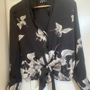 Svart skjorta med fågelmönster från ISTAY - Snygg svart skjorta från ISTAY i storlek M med unikt fågelmönster i vitt och beige. Skjortan har lång ärm och knyt framtill för en cool och trendig look. Materialet är mjukt och har en lätt glansig finish som känns lyxig.