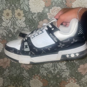 Vita och svarta sneakers från Vuitton - Säljer ett par snygga sneakers från Vuitton i vitt och svart med coola detaljer och mönster på sidorna. Skorna har snörning och en platt sula, perfekta för en trendig streetstyle-look.