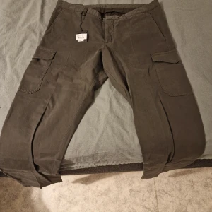 Peak Performance mörkgröna cargopants - Säljer ett par mörkgröna cargopants från Peak Performance i storlek W36/L34. Byxorna har flera praktiska fickor på sidorna och bak, klassisk cargomodell med rak passform och bälteshällor. Materialet är slitstark bomull som känns skönt mot huden.