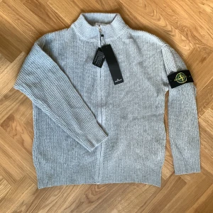 Grå stickad StoneIsland kofta - Säljer en grå stickad StoneIsland kofta med half zip och ribbad krage. Tröjan har långärmade ärmar och klassisk StoneIsland patch på vänster arm. Materialet är mjukt och grovstickat, perfekt för kyliga dagar. Snygg, stilren och tidlösa design som lyfter din outfit.