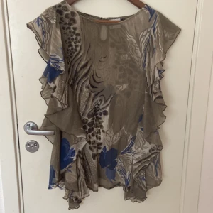 Mönstrad blus med volang från Gina - Säljer en luftig blus från Gina i storlek 44. Blusen har ett unikt mönster i beige, blått och brunt med volangdetaljer längs ärmar och sidor. Den är ärmlös och har en flowy passform, perfekt för dig som gillar statement-plagg med coola detaljer.