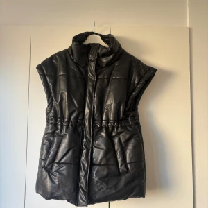 Svart dunväst från LXA - Svart dunväst från LXA med coolt quiltad design och hög krage. Västen har snörning i midjan för snygg passform och två praktiska fickor framtill. Materialet är skinnimitation som ger en edgy look. Perfekt att slänga över en hoodie eller tröja när det är kyligt ute.