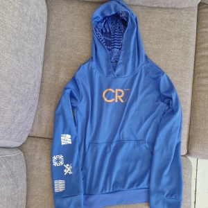 Blå CR7 hoodie från Nike - Snygg blå hoodie från Nike med CR7-logga i guld på bröstet och coola vita tryck på ena ärmen. Tröjan har huva med mönstrat foder och en stor magficka framtill. Tillverkad i mjuk polyester med lite elastan för extra komfort. Perfekt för dig som gillar sportig stil. Väldigt luftig och lätt klädsel som är perfekt för träning och vila i!