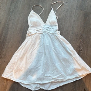 Vit omlottklänning från Zara - Supersöt vit kortklänning från Zara med omlottband i midjan och tunna axelband. Klänningen har v-ringad front, volangdetaljer vid midjan och är tillverkad i ett lätt, mjukt material som ger en flowy känsla. Perfekt för varma dagar och sommarhäng.