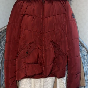 Mörkröd pufferjacka med fuskpäls - Snygg mörkröd pufferjacka med fluffig fuskpäls runt kragen. Jackan har dragkedja framtill, två fickor med knapp och quiltade detaljer. Perfekt för kalla dagar när du vill vara både varm och stilren.