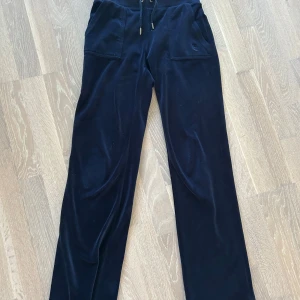 Mörkblå mjukisbyxor Juicy Couture - Snygga mörkblå mjukisbyxor från Juicy Couture i mjuk sammet. Byxorna har raka ben, snörning i midjan och fickor både fram och bak med silverknappar. Juicy Couture-logga broderad på ena fickan framtill. Perfekta för chill eller streetstyle.