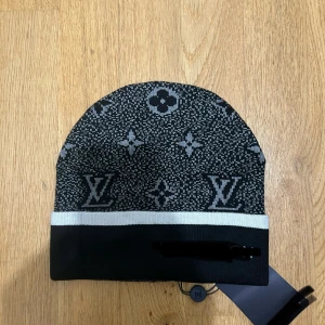 Svart mönstrig mössa  - Snygg svart mössa med klassiskt monogrammönster i grått och vit rand vid kanten. Perfekt för dig som gillar lyxiga accessoarer och vill sticka ut.