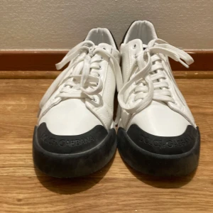 Dolce & Gabbana vita sneakers - Snygga vita sneakers från Dolce & Gabbana med svarta detaljer på tå och häl. Skorna har snörning och är tillverkade i skinn med en platt sula. Märkets logga syns både på häl och tå, samt på undersidan med ett mönstrat tryck. Perfekt för dig som gillar stilrena och exklusiva sneakers.