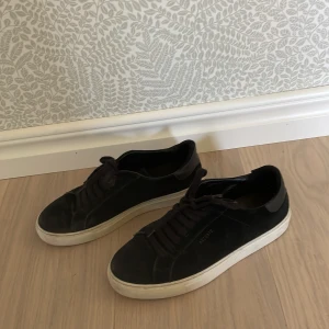 Svarta sneakers från Axel Arigato - Säljer ett par svarta sneakers från Axel Arigato med snygg, minimalistisk design. Skorna har svart mocka på ovansidan, svarta snören och en vit platt gummisula. Perfekt för dig som gillar stilrena och klassiska sneakers med en modern vibe.