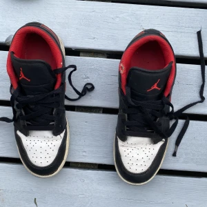 Nike Jordan 1 Low svart/röd 38 - Nike Jordan 1 Low sneakers i svart och vitt med röda detaljer och röd insida. Klassisk design med snörning, rund tå och platt sula. Swoosh-logga i rött på sidan och Jumpman på plösen. Skorna är tillverkade i skinn och har en stilren, sportig vibe.