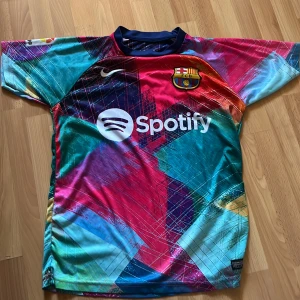 FC Barcelona Lewandowski Nike tröja - Säljer en FC Barcelona fotbollströja med Lewandowski och nummer 9 på ryggen. Tröjan har ett färgstarkt, abstrakt mönster i blått, rött, lila och turkos med vita detaljer. Officiell Nike-logga, Spotify sponsortryck och klubbmärke på bröstet. Kortärmad och tillverkad i lätt polyester.
