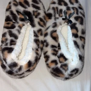 Leopardmönstrade fluffiga tofflor - Supermysiga tofflor med leopardmönster och fluffig pälsimitation. Insidan är vit och mjuk, perfekt för att hålla fötterna varma. Utsidan har bruna och svarta detaljer, och en liten svart rosett framtill. Sulan är beige med små prickar för extra grepp.