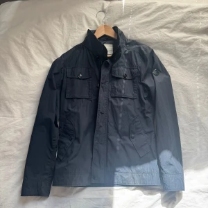 Mörkblå vindjacka från J.Lindeberg - Snygg mörkblå vindjacka från J.Lindeberg med hög krage, fyra fickor framtill och diskret logga på ärmen. Jackan har en clean look med knappar och dragkedja, perfekt för dig som gillar stilren design. Materialet är lätt och känns skönt under vår och höst.
