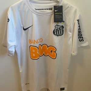 Santos Neymar 11 vit fotbollströja Nike - Snygg vit Santos fotbollströja från Nike med Neymar 11 på ryggen. Tröjan har korta ärmar, svart Nike-logga och Santos klubbmärke på bröstet. Materialet är lätt och ventilerande Dri-Fit polyester. Tryck i orange och svart, samt sponsorer på ärmar och bröst.