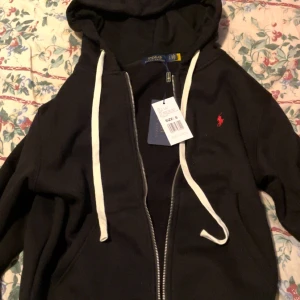 Svart hoodie från Polo Ralph Lauren - Svart hoodie från Polo Ralph Lauren med vit snörning och dragkedja. Klassisk röd logga broderad på bröstet. Tröjan har huva, känguruficka och är tillverkad i mjuk bomull. Perfekt för chill dagar och streetwear-stil.  