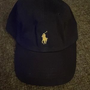 Mörkblå keps från Polo Ralph Lauren - Snygg mörkblå keps från Polo Ralph Lauren med klassisk gul broderad logga framtill. Ny utan prislapp fraktar inom 24 timmar. 