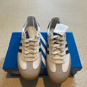 Adidas Samba sneakers vit/grön - Adidas Samba sneakers i klassisk stil med vit ovandel i skinn och mocka, gröna ränder på sidorna och gummisula. Snygga detaljer som snörning och rund tå. Perfekt för dig som gillar retrovibbar och sportig look. Kommer med originalkartong.