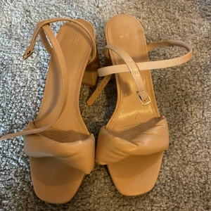 Beige sandaler med klack från H&M - Snygga beige sandaler från H&M med fyrkantig tå och bred, twistad rem framtill. De har smala ankelremmar med guldfärgat spänne och stabil blockklack. Tillverkade i syntetmaterial med mjuk finish, perfekta för dig som gillar stilrena och trendiga skor.
