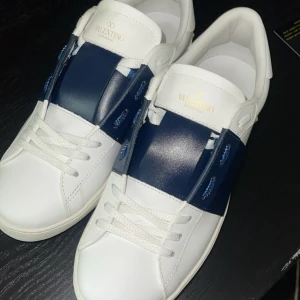 Valentino Garavani vita och blå sneakers - Snygga sneakers från Valentino Garavani i vitt skinn med en bred marinblå rem över mitten. Klassisk rund tå och vita snören. Perfekt för dig som gillar stilrena och exklusiva skor med en modern touch.
