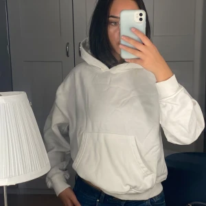 Vit hoodie med huva och ficka - Vit hoodie med klassisk huva och stor magficka. Tröjan har ribbade muddar vid ärmslut och nederkant, samt en loose passform som ger en chill vibe. Materialet är mjukt och känns som bomull, perfekt för att slänga på sig när man vill vara bekväm. Den har dock ett super litet hål som visas på bild