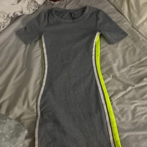 Grå ribbad klänning från H&M Divided - Säljer en grå ribbad klänning från H&M Divided i storlek XXS. Klänningen har korta ärmar och är figurnära med coola neongula och vita ränder längs sidorna. Materialet är mjukt och stretchigt, perfekt för dig som gillar sportig stil. Klänningen är i stretchigt material men jag säljer då den blev lite liten runt armarna. Pris kan diskuteras❤️🥰