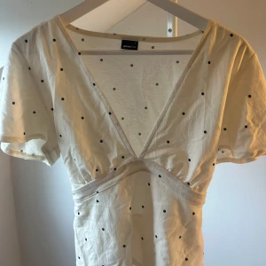 Prickig vit blus från Gina Tricot - Supersöt vit blus/topp från Gina Tricot med svarta prickar. Den har v-ringad hals, korta ärmar och markerad midja som ger en snygg siluett. Perfekt för sommarens alla tillfällen, gjord i mjuk bomull för skön känsla.