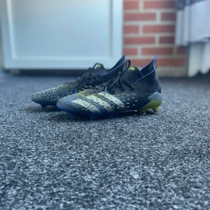 Adidas Predator fotbollsskor blå/gul - Säljer ett par Adidas Predator fotbollsskor med cool design i svart, blått och gult. Skorna har snörning, mönstrad ovandel med gula detaljer och en högre strumpliknande krage. Sulan är blå med gult och har dobbar för gräsplan. Perfekt för dig som vill sticka ut på planen.
