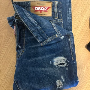 Blå slitna jeans från DSQUARED2 - Snygga blå jeans från DSQUARED2 med coola slitningar och trasiga detaljer på framsidan. Klassisk femficksmodell med tydliga kontrastsömmar och läderpatch bak. Perfekt för dig som gillar en edgy streetstil.