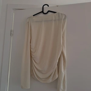 Krämvit långärmad topp från NA-KD - Figursydd långärmad topp från NA-KD i härlig glansig krämvit/ljusbeige färg. Väldigt sparsamt använd så den är i mycket bra skick! Strl M och normal i storleken. Gjord i mjukt och stretchigt material. Säljer då den tyvärr inte kommer till användning <33