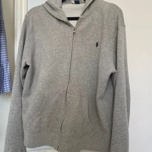 Grå hoodie från Polo Ralph Lauren - Den är i barn storlek men passar S. Klassisk grå hoodie från Polo Ralph Lauren med dragkedja och huva. Tröjan har en liten broderad logga på bröstet och är tillverkad i mjuk bomull. Perfekt för en avslappnad stil och enkel att matcha med andra plagg. De finns en fläck se sista bilden. 