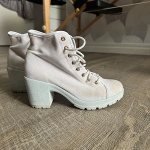 Vita boots med snörning och klack - Säljer ett par vita boots med grov klack och snörning framtill. Skorna har rund tå och är tillverkade i ett tygmaterial med robust sula. Lite missfärgningar