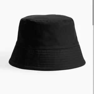 Svart bucket hat - En klassisk svart bucket hat med rund form och nedvikt brätte. Hatten är tillverkad i ett slätt material och passar perfekt för en stilren look. Enkel att matcha med olika outfits och skyddar mot solen.