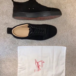 Svarta sneakers från Christian Louboutin - Säljer ett par svarta sneakers från Christian Louboutin med klassisk röd sula och broderad logga på hälen. Skorna har snörning och är tillverkade i mocka med svarta detaljer. Perfekta för dig som vill ha något extra till din outfit.