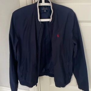 Marinblå bomberjacka från Polo Ralph Lauren - Säljer en stilren marinblå bomberjacka från Polo Ralph Lauren med röd broderad logga på bröstet. Jackan har dragkedja framtill, ribbade muddar och klassisk krage med vit kant. Perfekt för dig som gillar tidlös och sportig stil. Hör av er vid frågor.