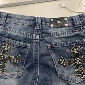 Blå jeans med broderade kors och strasstenar från Inzim - Unika blå jeans med två stora broderade kors i svart, grönt och guld på bakfickorna, dekorerade med strasstenar. Klassisk femficksmodell med silverfärgad metallpatch i midjan. Perfekt för dig som vill sticka ut.