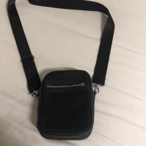 Svart axelväska med dragkedja - En stilren svart axelväska med justerbart band och en praktisk dragkedja framtill. Väskan har en rektangulär form och är perfekt för att bära med sig det viktigaste. Passar dig som vill ha en enkel och snygg accessoar till vardagen.