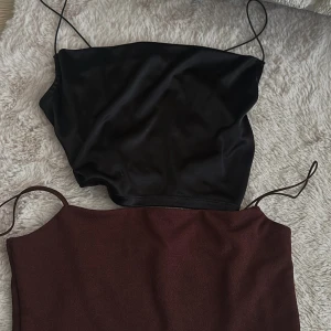 Svart och brun croppad topp med tunna axelband - Två stilrena croppade toppar, en i svart satin och en brun, båda med tunna axelband. Perfekta att matcha med jeans eller kjol för en trendig look. Den bruna är storlek M och den svarta S