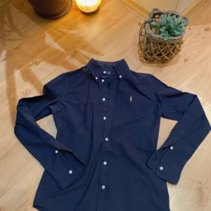 Mörkblå skjorta från Ralph Lauren - Säljer en klassisk mörkblå skjorta från Ralph Lauren i modellen Custom Fit. Skjortan har långa ärmar, knappar framtill och den ikoniska broderade loggan på bröstet. Perfekt för dig som gillar stilrena plagg med snygg passform.