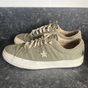 Olivgröna Converse sneakers med stjärndetalj - Säljer ett par olivgröna Converse sneakers med vit sula och snörning. Skorna har en tydlig stjärndetalj på sidan och är tillverkade i canvasmaterial. Det som ser ut att vara slitningar är en del av designen och det finns inga större defekter på skon förutom lite heel drag därav låga priset! Hör ev er vid frågor! 
