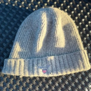 Grå ribbad mössa från GANT - Säljer en stilren grå ribbstickad mössa från GANT med uppvikt kant och liten broderad logotyp framtill. Perfekt för kyliga dagar och enkel att matcha med olika outfits. 6/10 på skick perfekt på kalla sommar kvällar