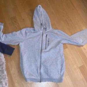 Grå hoodie med dragkedja - Säljer en ljusgrå hoodie med huva och dragkedja framtill. Jackan har långa ärmar och en enkel, stilren design. Perfekt för en avslappnad look.