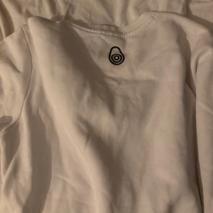 Vit sweatshirt från Sail Racing - Vit sweatshirt från Sail Racing med svart logotyp tryckt över bröstet. Tröjan har rund halsringning med svart och vit randig detalj på kragen. Klassisk och stilren design, perfekt för en sportig look.den är i smål i barn storlek.