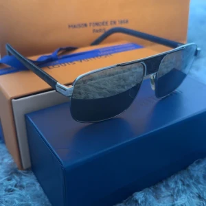 Louis Vuitton solglasögon med svarta glas - Snygga solglasögon från Louis Vuitton med rektangulära svarta glas och silvriga detaljer. Skalmarna är svarta med diskret LV-mönster i vitt. Perfekt accessoar för dig som vill sticka ut med stil. 6/10 skick.