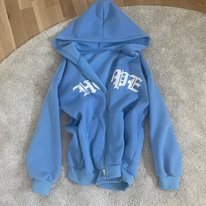 Blå hoodie med tryck - Säljer en ljusblå hoodie med huva och dragkedja. På bröstet står det 'HOPE' i vit gotisk stil. Tröjan har ribbade muddar och fickor framtill. Perfekt för en chill och avslappnad stil.