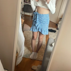 Blå omlottkjol med volang och blommigt mönster - Säljer en söt blå omlottkjol med vitt blommigt mönster och volangkant. Kjolen knyts i sidan och har en kort längd och dragkedja bak. Passar perfekt till varmare dagar. Superfin och trendig modell! Sen säljer jag också en grön kjol som är ganska liknande modell fast stretch i midjan. Köp båda för 150 eller en för 90.