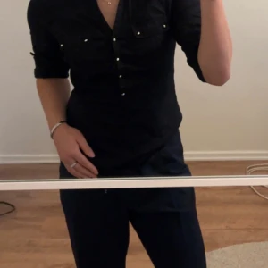 Svart skjorta med fina detaljer - Svart skjorta med knapp-detaljer i guldfärg. Har vikt upp ärmarna, men man kan vika ut dem så det blir en hel armlängd. Använd, men inga tecken på slitage. Storlek M, men passar även S