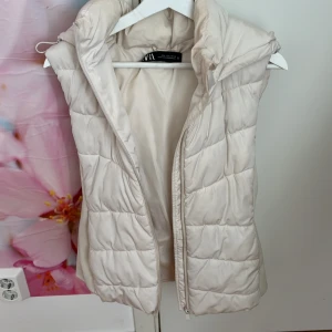 Vit dunväst från Zara - Säljer en vit dunväst från Zara med hög krage och huva. Den är i storlek L men kan användas för någon som är storlek M samt S.  Västen har quiltad design och stängs med dragkedja framtill. Perfekt för lager-på-lager och passar till många olika stilar.