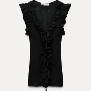 Svart volangprydd ribbad topp från Zara - Säljer en svart ärmlös topp från Zara med djup v-ringning och volangdetaljer längs axlar och fram. Toppen har knyt framtill och är ribbad i materialet för en snygg passform.